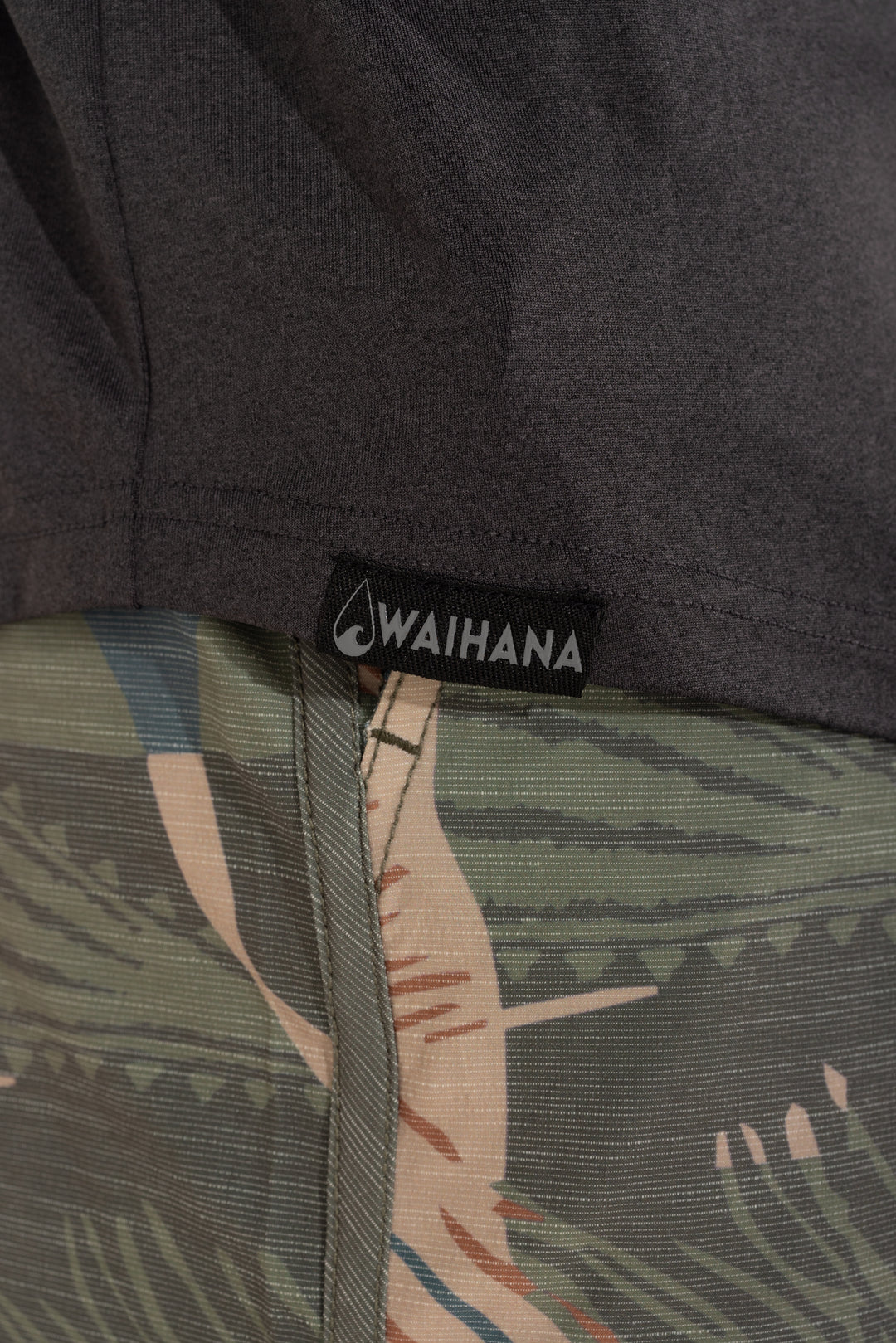 Close-up of the Waihana woven logo label on the hem of a charcoal Mariner hooded long sleeve sun shirt, layered over tropical-print shorts.