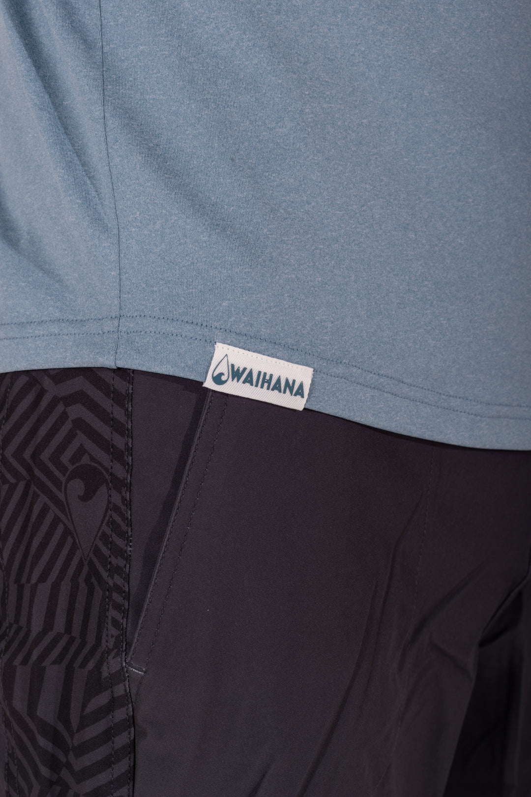 Close-up of the Waihana woven logo label at the hem of a Mariner Blue hooded long sleeve sun shirt, paired with black patterned board shorts.