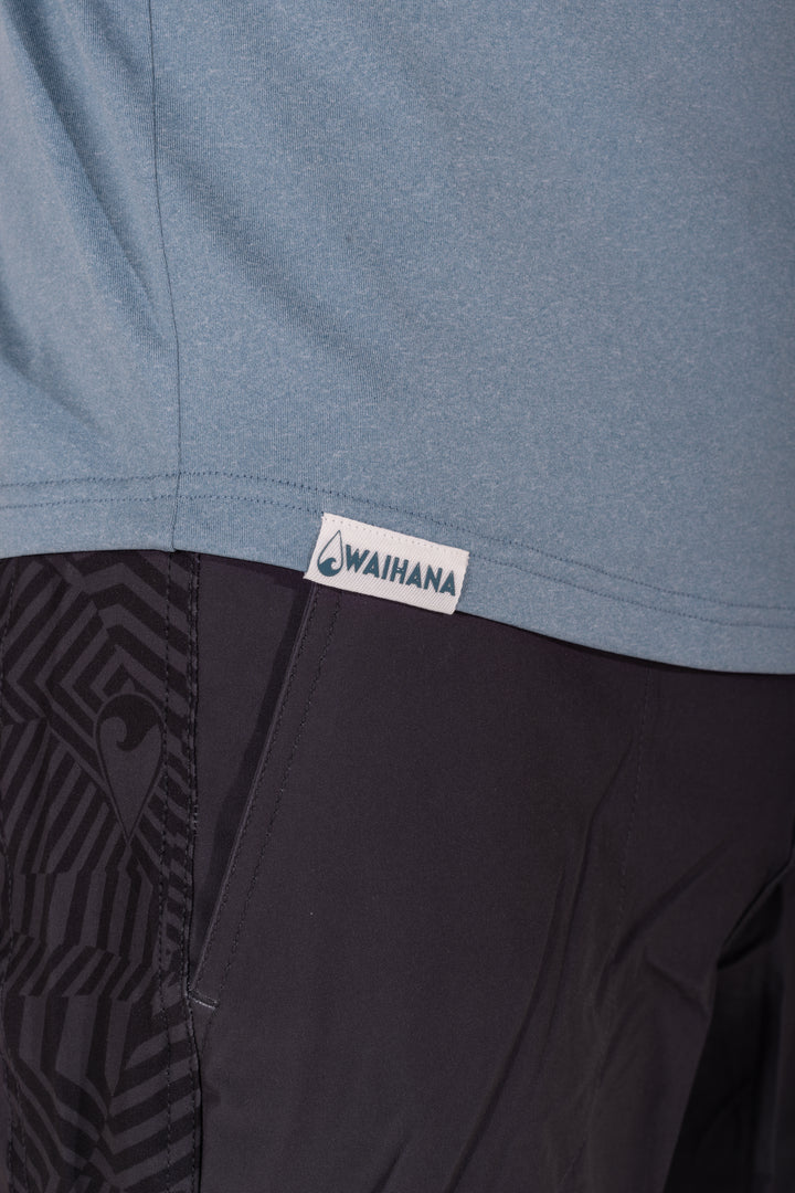Close-up of the Waihana woven logo label at the hem of a Mariner Blue hooded long sleeve sun shirt, paired with black patterned board shorts.